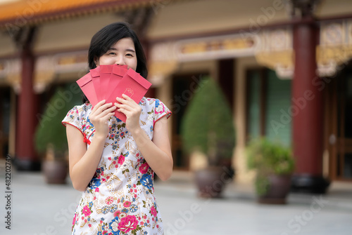 Pretty Asia woman wearing Chinese traditional dress cheongsam or qipao holding red envelop and kiss. Standing in the temple with copy space. Feeling happy on new year celebration.