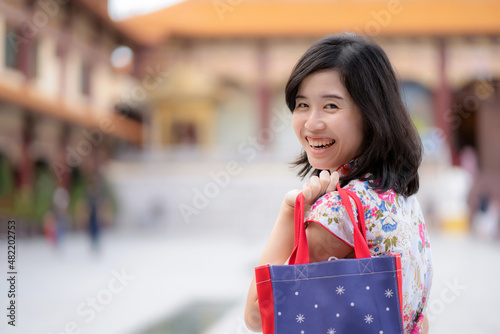 Confident Asia woman wearing Chinese traditional dress cheongsam or qipao standing with blur background. Happy on Chinese lunar new year celebration with smile. Holding shopping bag with copy space