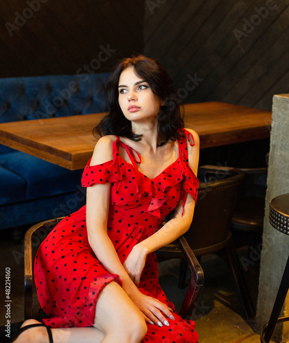 Sitting young dark haired woman with white skin in dark wooden room in bright red dress