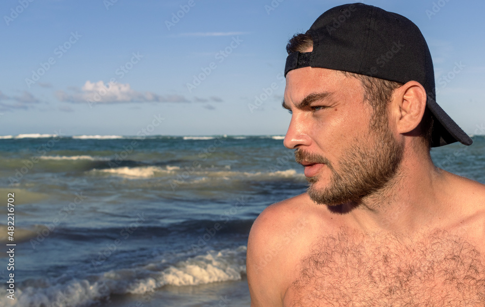 Handsome confident individual shirtless man in the cap standing on the beach and enjoying the nature, traveler man side portrait. Handsome confident individual shirtless man in the cap standing on the beach and enjoying the nature, traveler man side portrait.