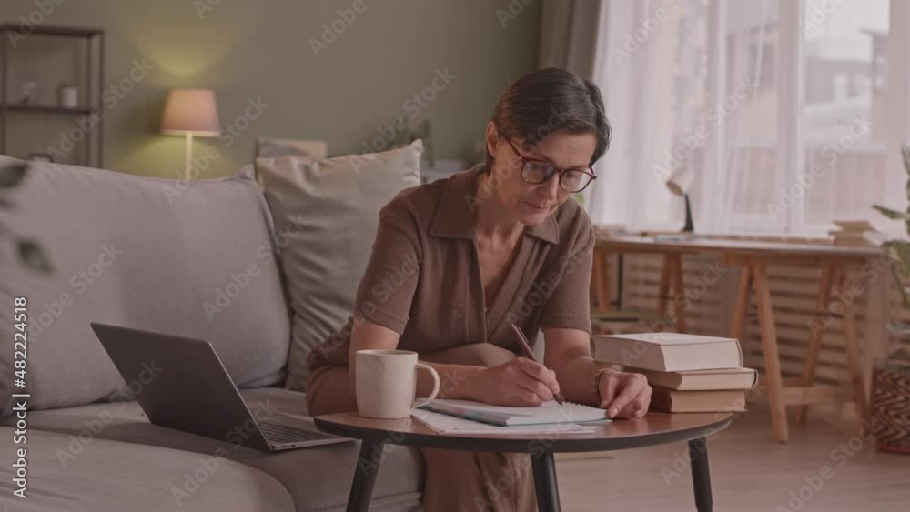 Medium slowmo shot of 50-year-old Caucasian woman studying online from home, watching video lesson on laptop and taking notes in copybook Medium slowmo shot of 50-year-old Caucasian woman studying online from home, watching video lesson on laptop and taking notes in copybook