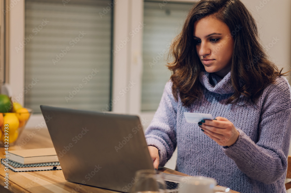 Hispanic woman with laptop using credit card in kitchen at home Hispanic woman with laptop using credit card in kitchen at home