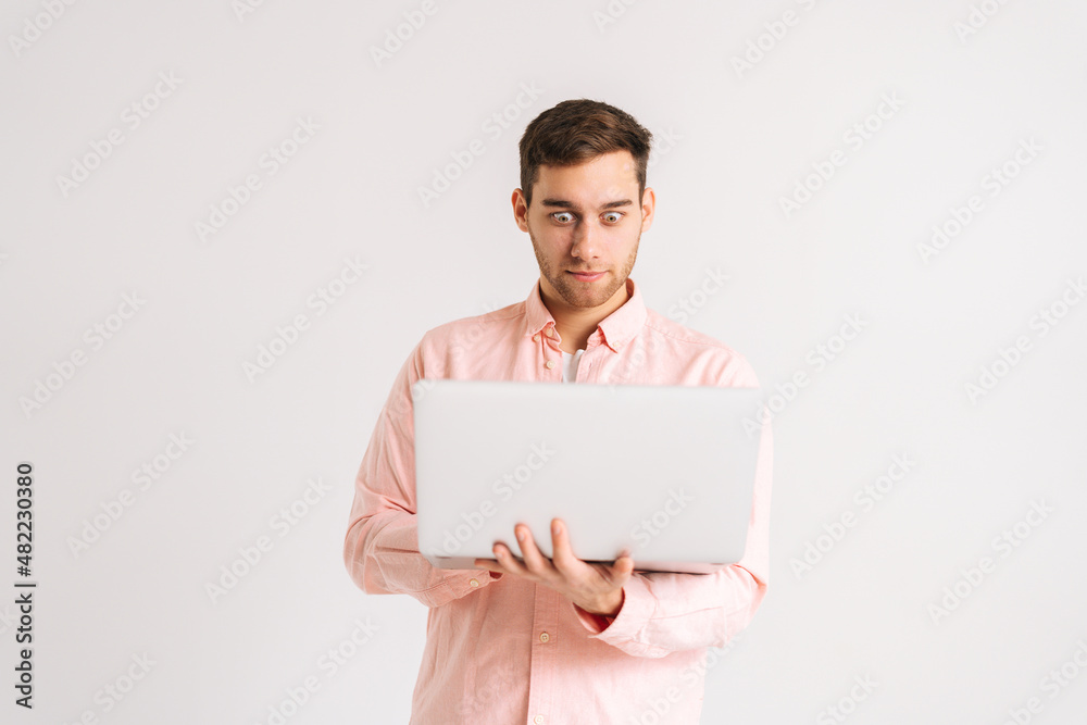 Portrait of shocked young man standing with laptop looking on screen with confused puzzled face on white isolated background. Studio shot of pensive male posing with pc looking at screen