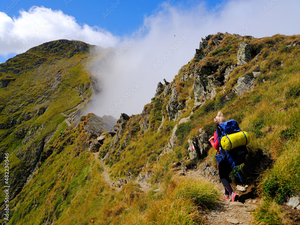 Young woman with a backpack descends the rocky terrain with a blurred view of the mighty green mountains and the blue sky with clouds. Fagaras Mountains, Carpathians, Romania