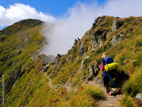 Fotografie Young woman with a backpack descends the rocky terrain with a blurred view of the mighty green mountains and the blue sky with clouds