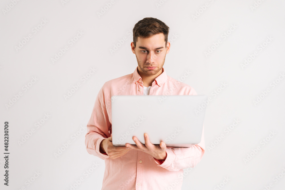 Portrait of shocked young man standing with laptop looking on screen with confused puzzled face on white isolated background. Studio shot of pensive male posing with pc looking at screen Portrait of shocked young man standing with laptop looking on screen with confused puzzled face on white isolated background. Studio shot of pensive male posing with pc looking at screen