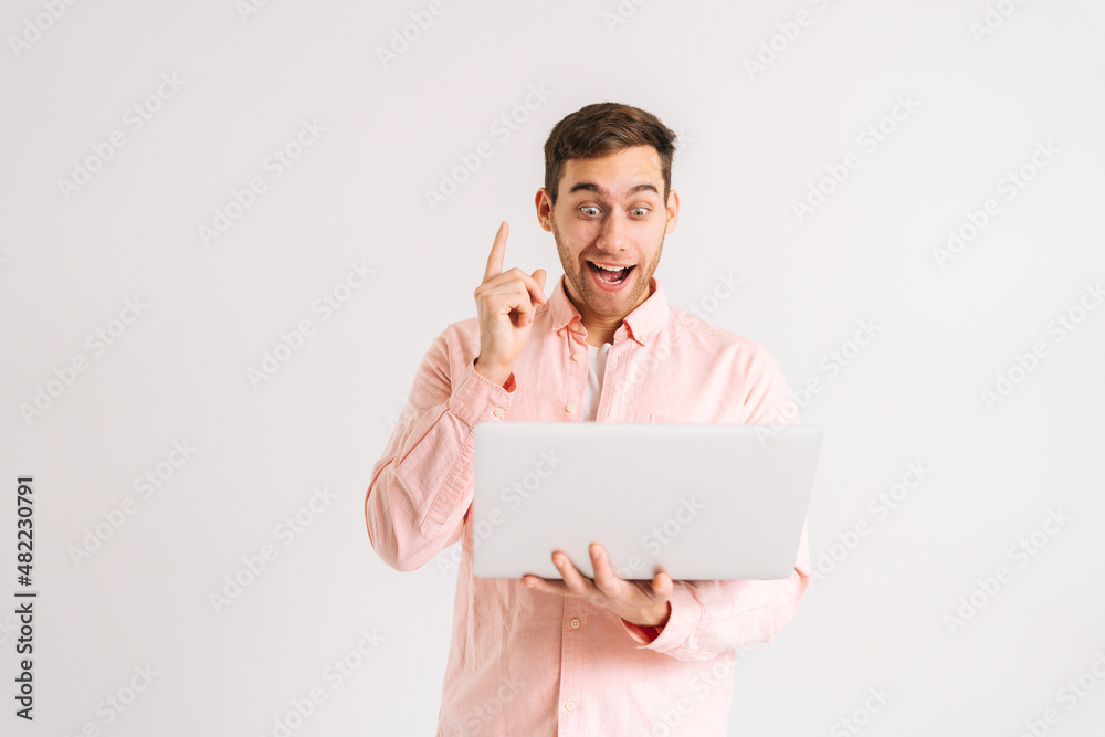 Portrait of amazed excited young man standing working on laptop computer pointing index finger up with happy face on white isolated background. Studio shot of happy male student posing with notebook. Portrait of amazed excited young man standing working on laptop computer pointing index finger up with happy face on white isolated background. Studio shot of happy male student posing with notebook.