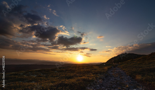 Mountain sunset with scattered clouds. A rocky path leads to a human with a dog