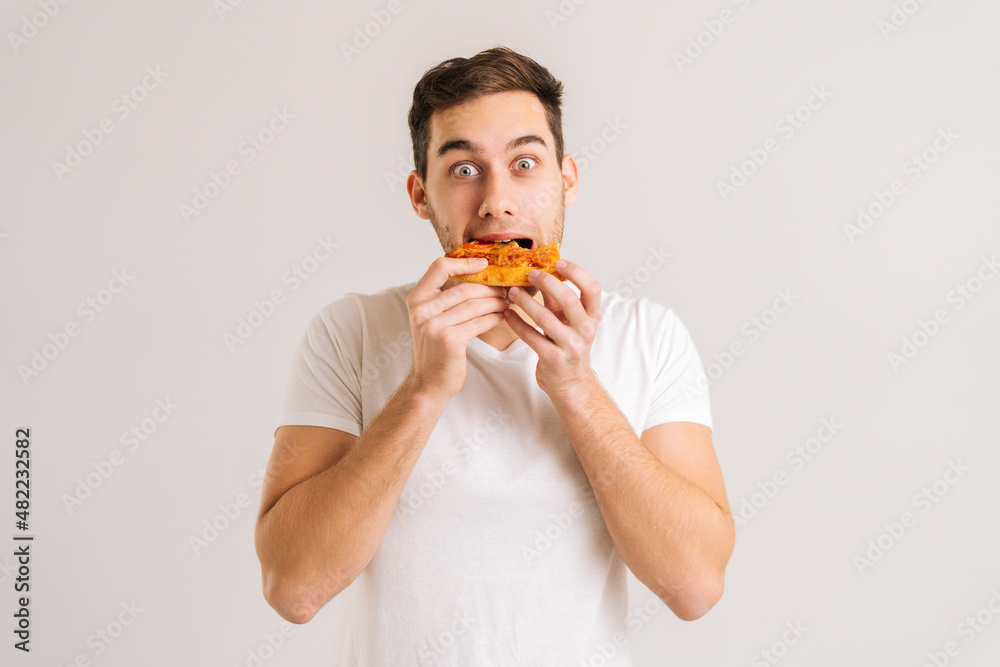 Portrait of exctited young man bitting delicious slice of pizza looking at camera on white isolated background. Studio shot of handsome male student eating tasty food. Portrait of exctited young man bitting delicious slice of pizza looking at camera on white isolated background. Studio shot of handsome male student eating tasty food.