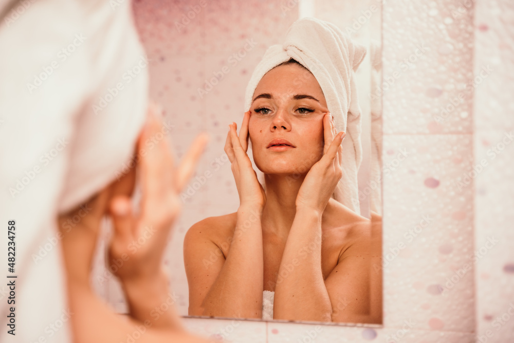 Young beautiful woman using face cream moisturizing lotion after bath. Pretty attractive girl in a towel on her head stands in front of a mirror in a home bathroom. Daily hygiene and skin care Young beautiful woman using face cream moisturizing lotion after bath. Pretty attractive girl in a towel on her head stands in front of a mirror in a home bathroom. Daily hygiene and skin care