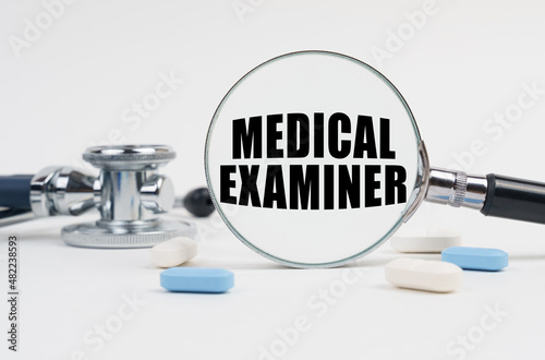 On a white surface lie pills, a stethoscope and a magnifying glass with the inscription - MEDICAL EXAMINER photo