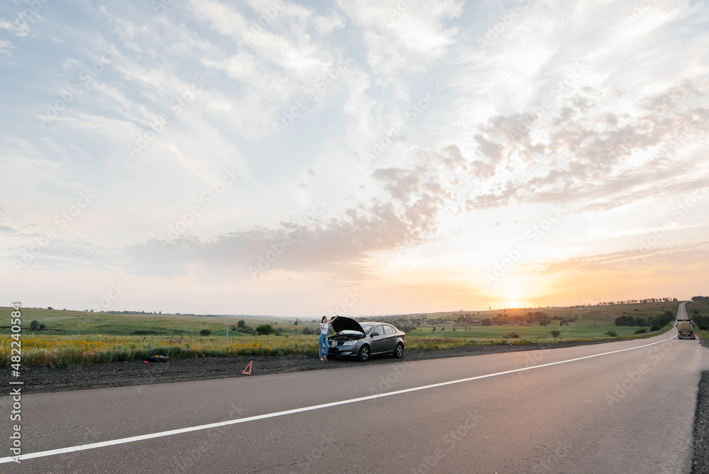 A young girl stands near a broken-down car in the middle of the highway during sunset and tries to call for help on the phone. Waiting for help. Car service. Car breakdown on road.