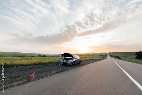 A young girl stands near a broken car in the middle of the highway during sunset and tries to repair it. Troubleshooting the problem. Waiting for help. Car service. Car breakdown on road..