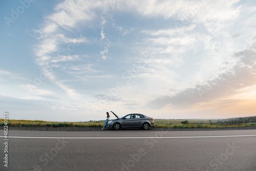 A young girl stands near a broken-down car in the middle of the highway during sunset and tries to call for help on the phone. Waiting for help. Car service. Car breakdown on road. © Andrii