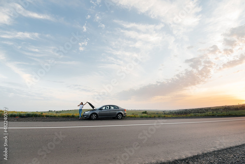 A young girl stands near a broken-down car in the middle of the highway during sunset and tries to call for help on the phone. Waiting for help. Car service. Car breakdown on road.