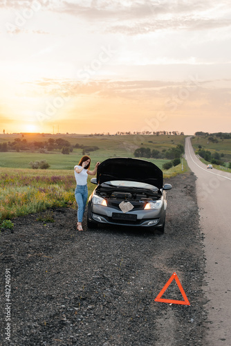 A young girl stands near a broken car in the middle of the highway during sunset and tries to call for help on the phone and start the car. Waiting for help. Car service. Car breakdown on the road.