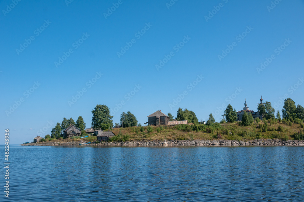 View of Church of Nativity of Virgin and old wooden sheds on shore of Lake Segozero in summer, Karelia, Russia