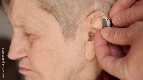 Close-up view of hands inserts hearing aid to the elderly woman's ear. Senior woman with symptoms of hearing loss wears a hearing aid device to hear properly. 
 photo