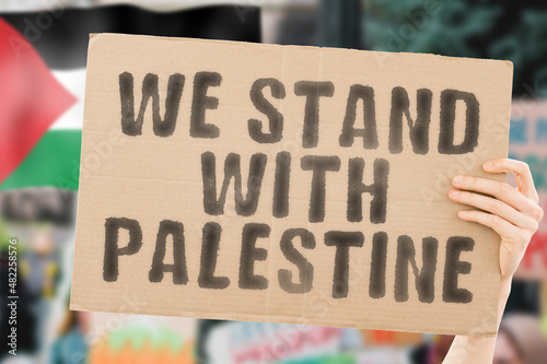 The phrase " We stand with Palestine " on a banner in men's hand with blurred Palestinian flag on the background. Support. Patriot. Community. Free. Arabian. Protest. Nation. Demonstration. Arabic