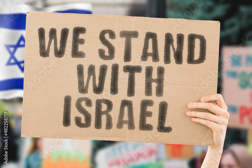 The phrase " We stand with Israel " on a banner in men's hand with blurred Israeli flag on the background. Support. Patriot. Protect. Free. Jewish. Protest. Nation. Demonstration. Protection