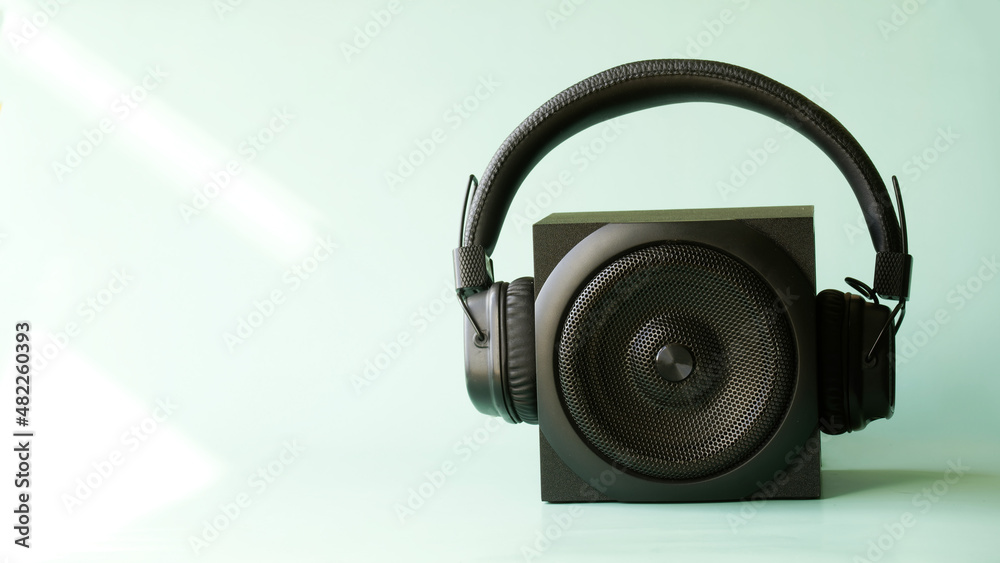 Black audio speaker and large wireless headphones on a pastel blue-green background illuminated by the rays of the sun. The concept of studio audio equipment. Copy space for an inscription Black audio speaker and large wireless headphones on a pastel blue-green background illuminated by the rays of the sun. The concept of studio audio equipment. Copy space for an inscription
