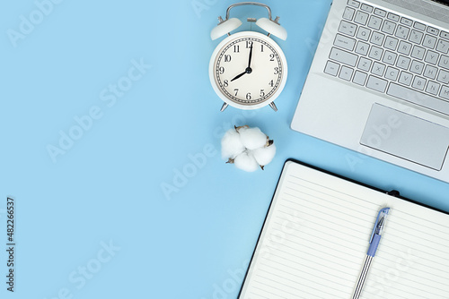 flat lay of a laptop, notebook, pen, alarm clock on blue background with copy space