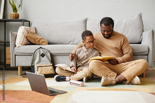 Full length portrait of African-American father and son sitting on floor while doing homework together, copy space