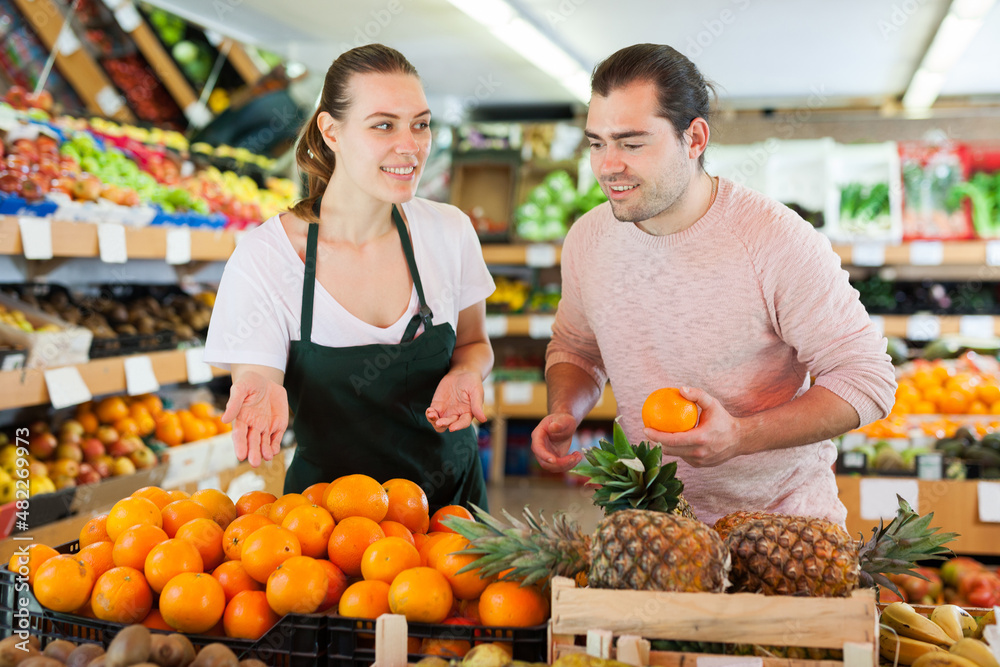 Young woman wearing apron selling fresh oranges to man customer in fruit store