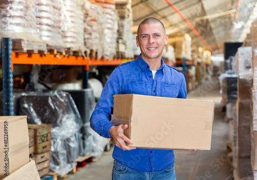Caucasian man worker holding cardboard box while standing in storehouse, looking at camera and smiling.