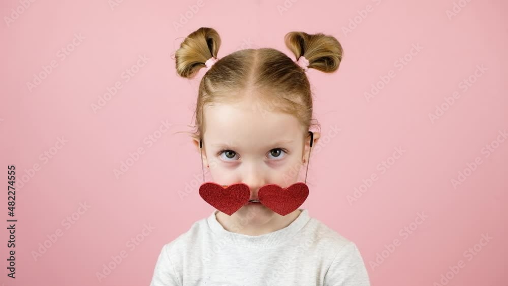 Funny little blonde girl smiling and playing with red heart shape sunglasses on pink background. Valentines Day or Mothers Day concept