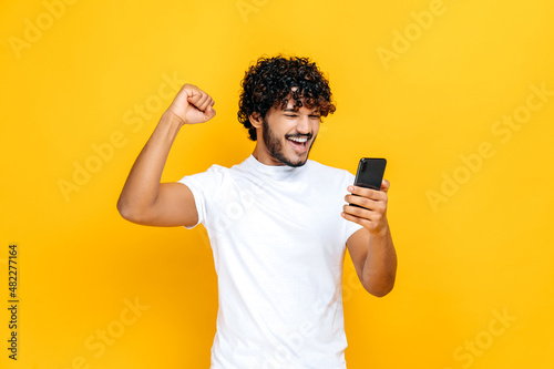 Amazed joyful excited Indian or Arabian guy holds smartphone, get unexpected news, winning lottery, stands on isolated orange background, cheerful facial expression, toothy smile, gesturing with fist
