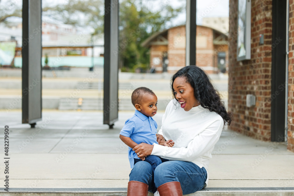 A beautiful African-American mom sitting on steps outdoors and hugging her toddler age son and they are both smiling