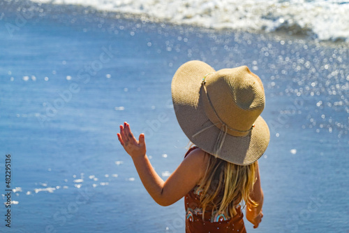 Young child, girl on beach with large brimmed hat sun and swim suit standing next to the ocean water on holiday photo