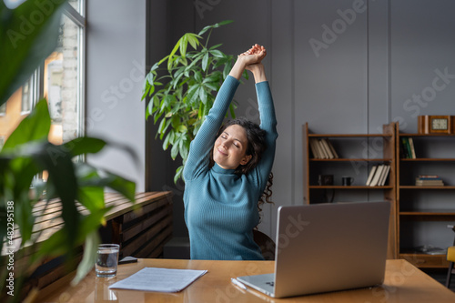 Happy businesswoman warming up body and muscles at workplace, feeling satisfied with work done, smiling female employee resting from computer screen. Well-being, productivity and happiness at work photo