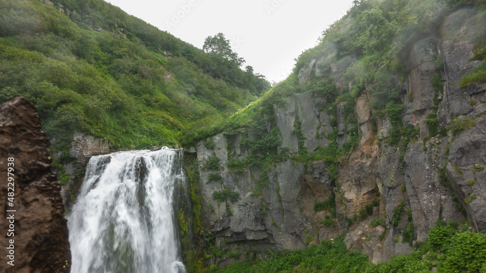 The waterfall flows from the sheer cliff. Water is foaming. On the slope of the mountain green vegetation. Kamchatka.