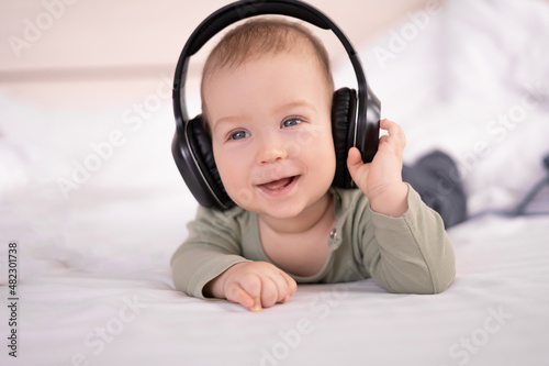 portrait of baby boy 4 months old with headphones on a white background, the baby listens to music with an earphone, a newborn on the bed smiles looks