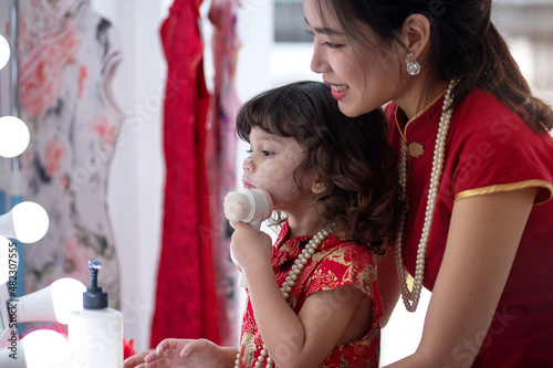 Happy mother with her lovely daughter wearing a traditional Chinese red dress preparing to celebrate the Chinese New Year, doing hairstyles and makeup, in front of mirror with light bulbs