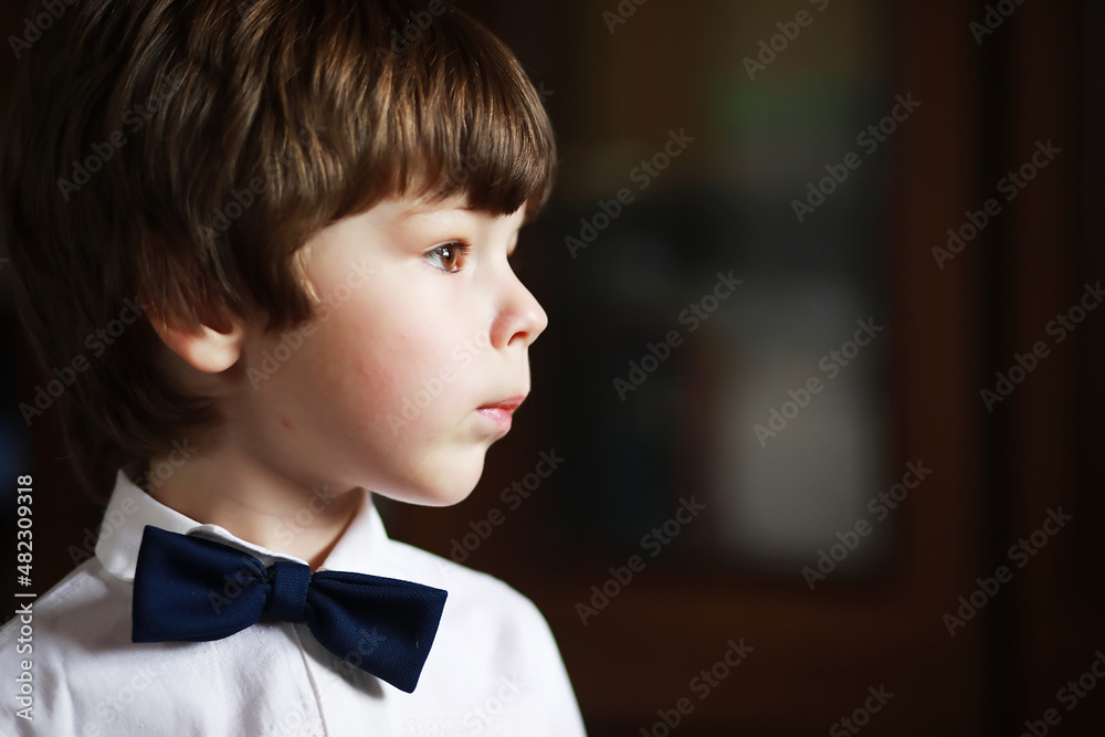 Portrait of a boy with brown eyes in a shirt and a black bow tie on a dark background. The child is emotional, smiling, happy. Portrait of a boy with brown eyes in a shirt and a black bow tie on a dark background. The child is emotional, smiling, happy.