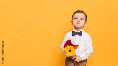 A boy in a white shirt, trousers and bow tie on a yellow background holds a bouquet of gerberas. A gift for my mother, grandmother. With space for your text.