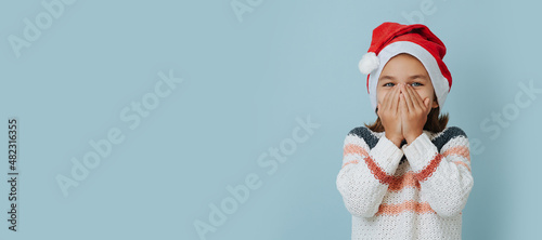 Laughing little girl in christmas hat covering her face over blue