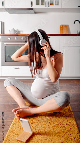 Pregnant doing workout on orange fitness mat. Pragnant yoga at home in the kitchen. Pregnant in white headphones.