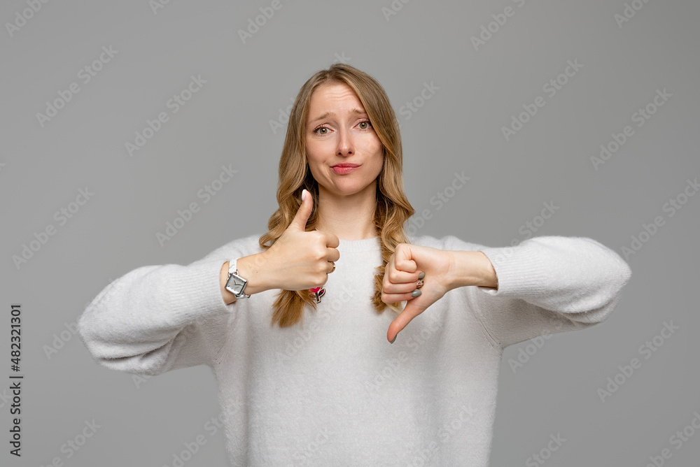 Fototapeta premium Puzzled blonde girl showing thumb up and down. Skeptical thinking woman judging deciding how to rate a situation, wears white sweater, gray background