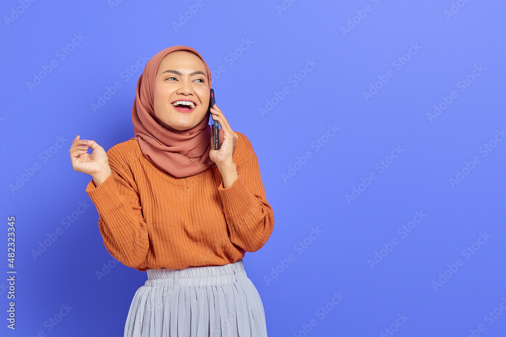 Cheerful beautiful Asian woman in brown sweater and hijab talking on mobile phone while looking aside isolated over purple background. People islam religious concept Cheerful beautiful Asian woman in brown sweater and hijab talking on mobile phone while looking aside isolated over purple background. People islam religious concept