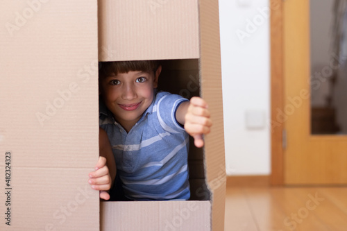 Smiling little boy playing in new house. Schoolboy with dark hair hiding in big cardboard box. Real estate, purchase, family concept