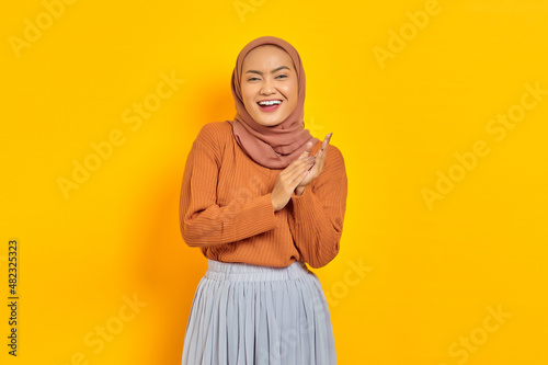 Beautiful smiling Asian woman in brown sweater and hijab rubs hands feels joyful has white teeth, looking at camera with confident isolated over yellow background. People islam religious concept © Bangun Stock Photo