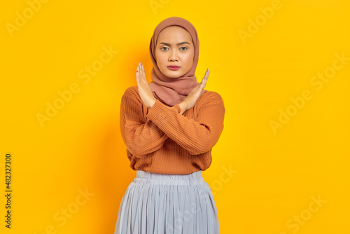 Beautiful annoyed Asian woman in brown sweater and hijab crossing hands makes stop gesture, demonstrates rejection isolated over yellow background. People islam religious concept © Bangun Stock Photo