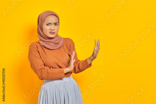 Beautiful annoyed asian woman in brown sweater and hijab makes stop gesture, demonstrates rejection isolated over yellow background. people religious lifestyle concept © Bangun Stock Photo
