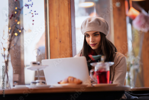 Smiling young woman wearing beret had and scarf while using her laptop at the cafe