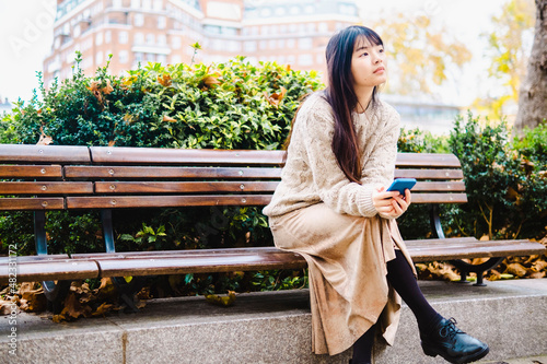 Thoughtful woman sitting with smart phone on bench at park photo