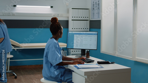 Medical assistant typing on computer keyboard in cabinet. Woman working as nurse with uniform using monitor for checkup appointments and healthcare practice while sitting as desk.